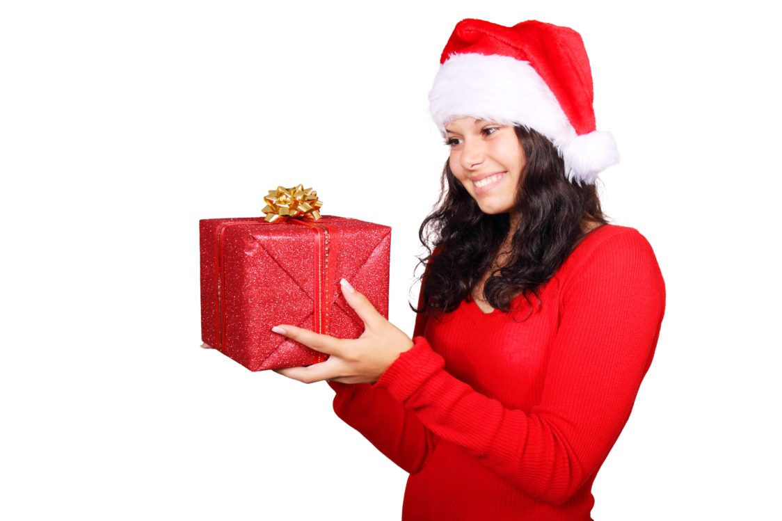 Una ragazza vestita in rosso sorridente tiene in mano un pacco regalo incartato con nastro e carta natalizia. Regali di Natale ecologici e sostenibili, i consigli di NaturalMania.it